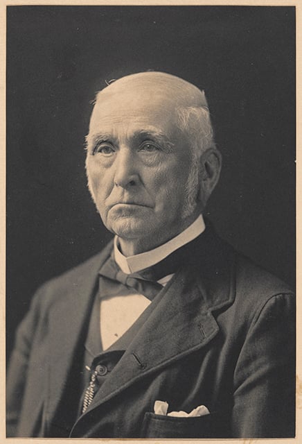 Black-and-white photograph of Joel Bean Bassett in his seventies. He is wearing a three-piece suit and bowtie and frowning, looking past the photographer to the left side of the image. 
